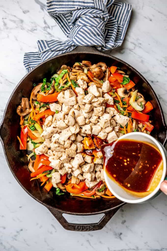 Sauce being poured over cubed chicken on a pile of stir fried vegetables in a cast iron skillet.