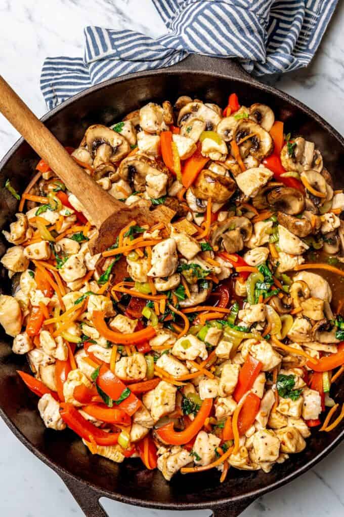 A wooden spoon scooping up some chicken and veggie stir fry from a cast iron skillet.