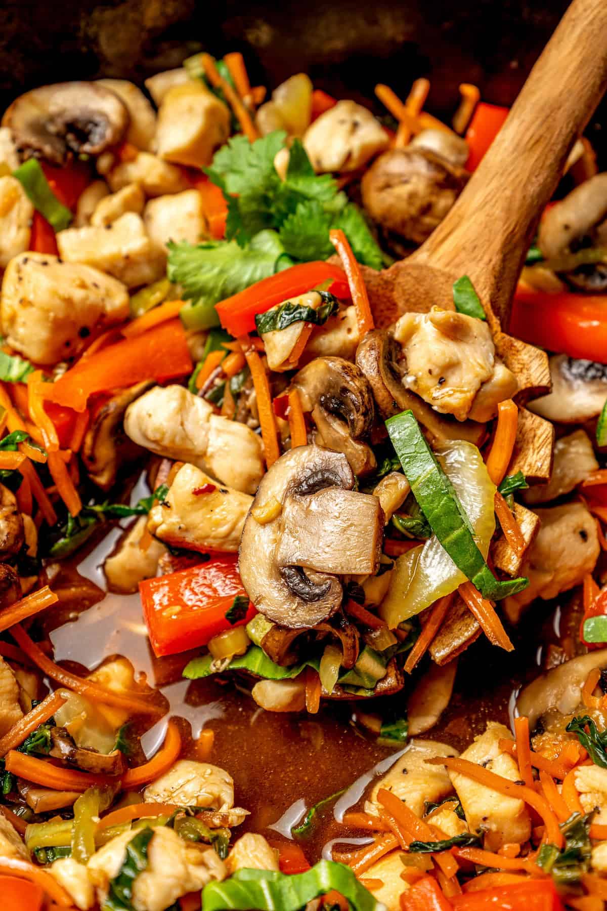 Close up of a wooden spoon scooping up some chicken and veggie stir fry from a cast iron skillet.