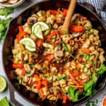 A wooden spoon resting in a chicken and veggie stir fry mix in a cast iron skillet with a text overlay on the photo.