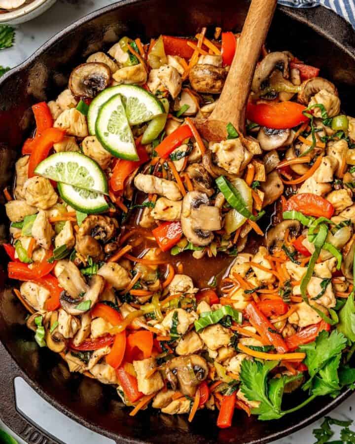 A wooden spoon with a scoop of vegetables in a cast iron skillet full of a chicken stir fry.