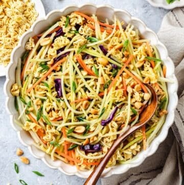 Close up of a wooden spoon resting in a large bowl of Asian style coleslaw with peanuts and ramen.