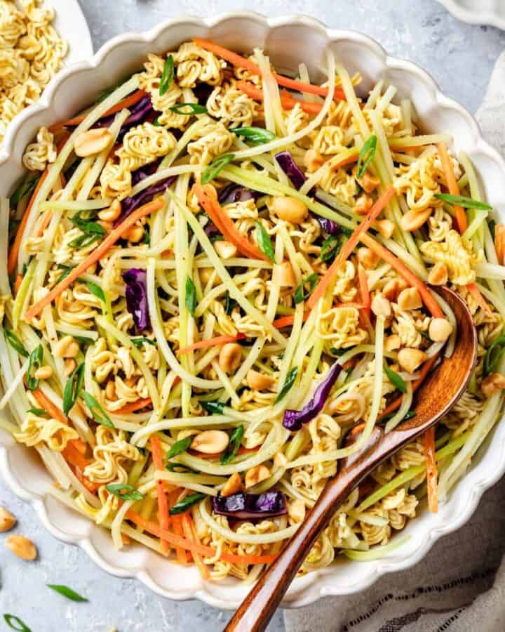 Close up of a wooden spoon resting in a large bowl of Asian style coleslaw with peanuts and ramen.