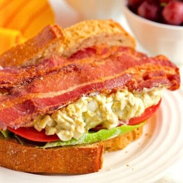 Close up of an egg salad sandwich with bacon, lettuce and tomato on thick bakery bread with a bowl of red grapes in the background.