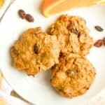 Three oatmeal raisin breakfast cookies on a small white plate with raisins scattered around and an orange slice to the side.