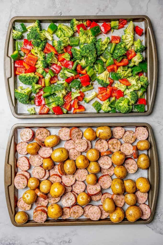 Two sheet pans, one with smoked sausage and baby potatoes, the other with peppers and broccoli, before roasting.