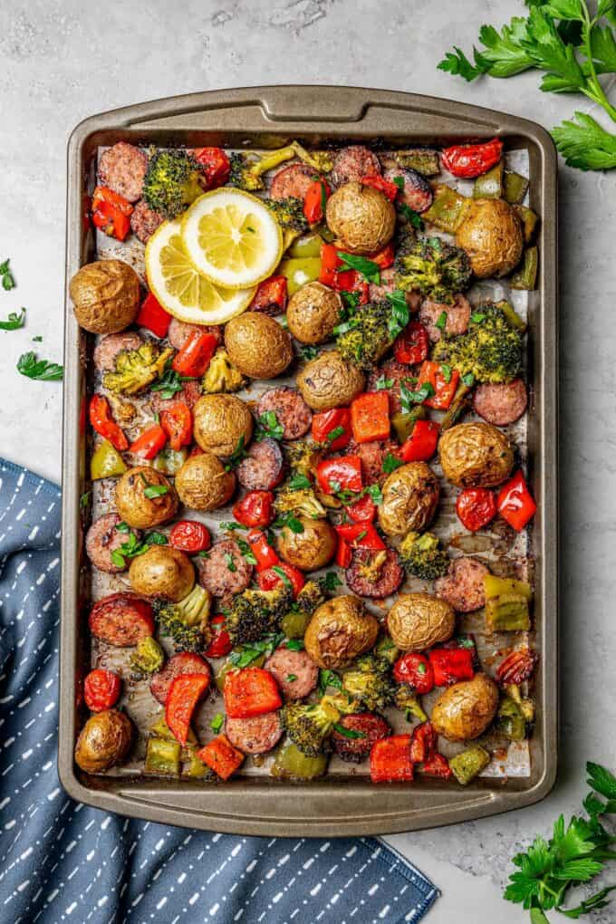 A sheet pan with roasted smoked sausage, potatoes, peppers, broccoli and tomatoes with lemon slices for garnish.