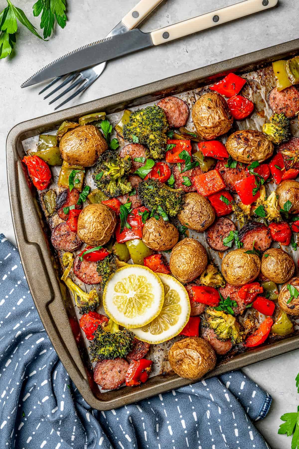 A sheet pan with roasted potatoes, veggies and sausage with utensils to the side.