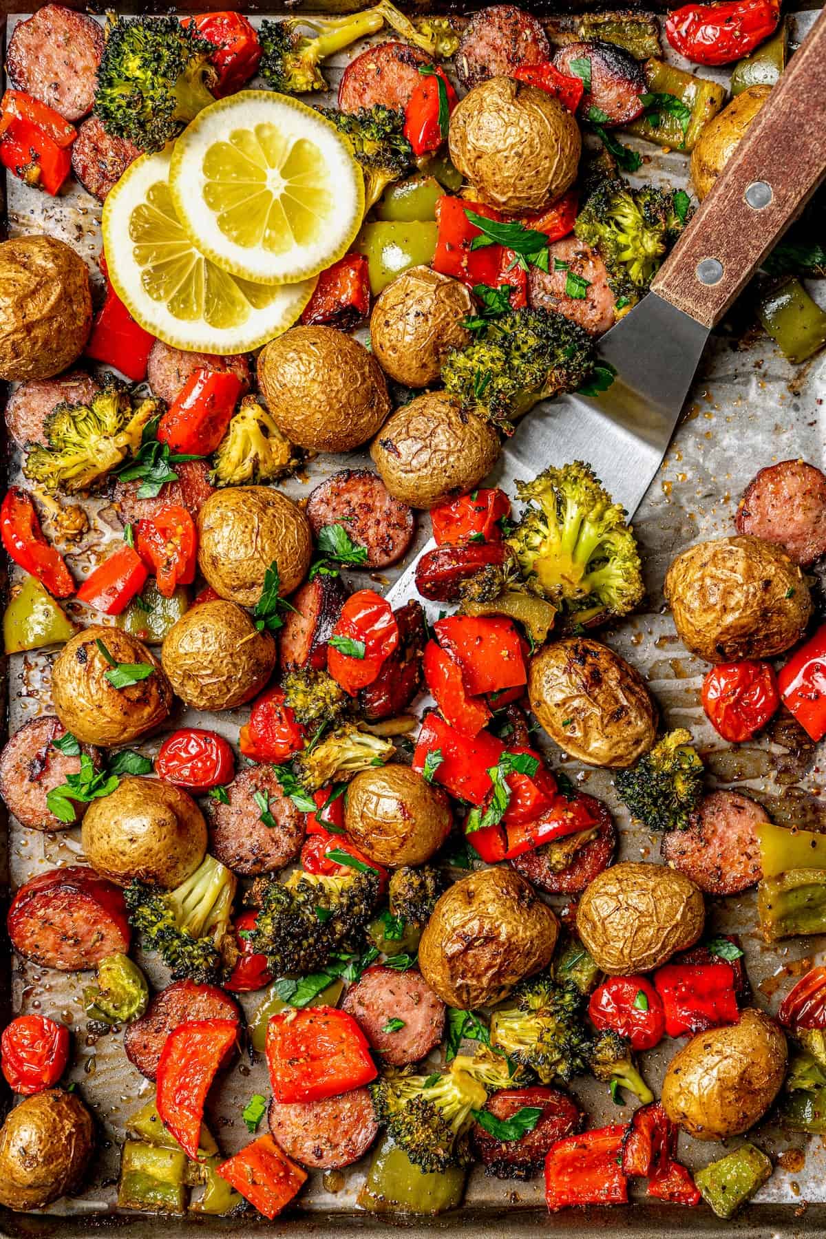 A spatula scooping up some roasted veggies and sausage from a sheet pan dinner.