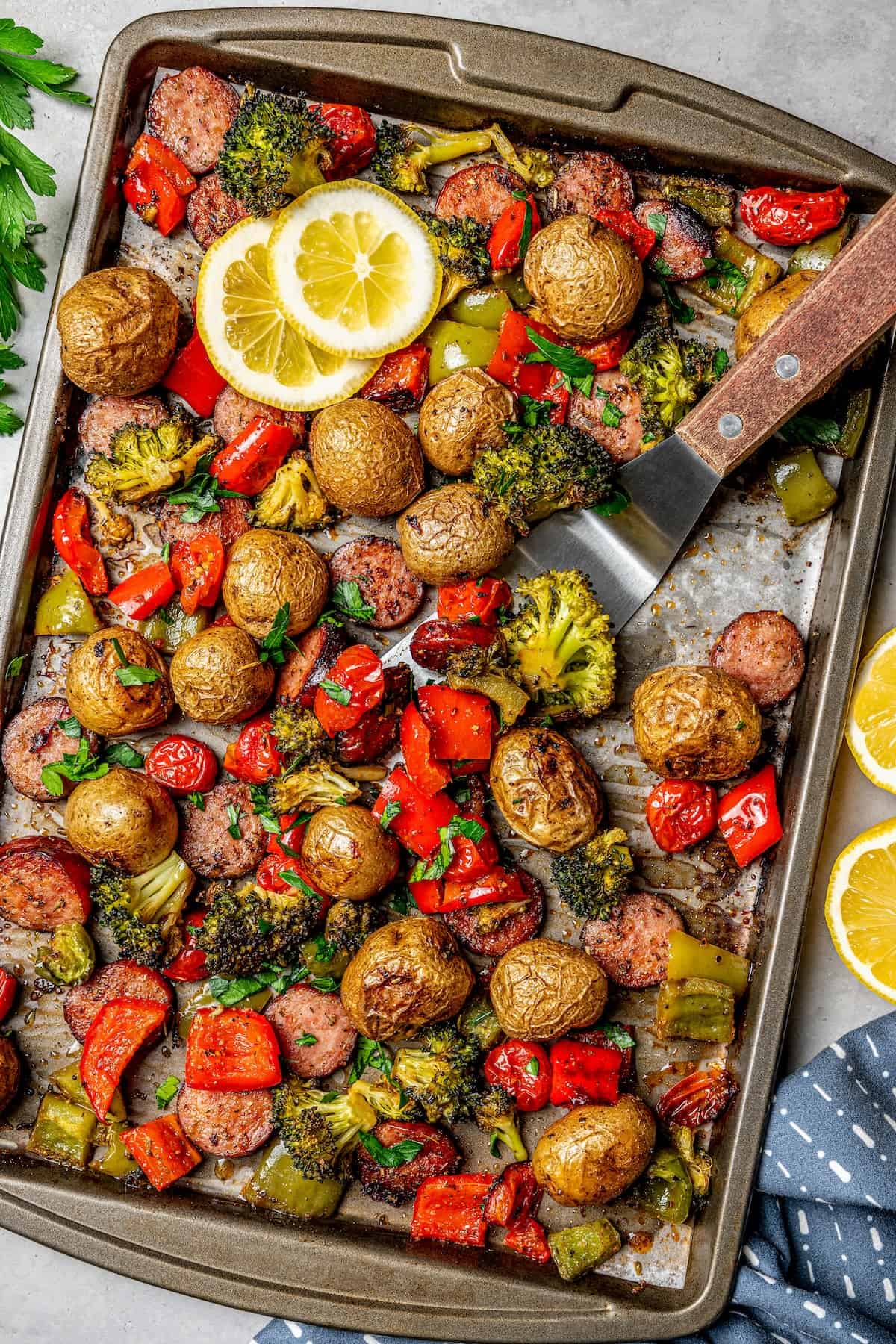 A spatula scooping up some roasted peppers, potatoes, broccoli and sausage from a sheet pan dinner.