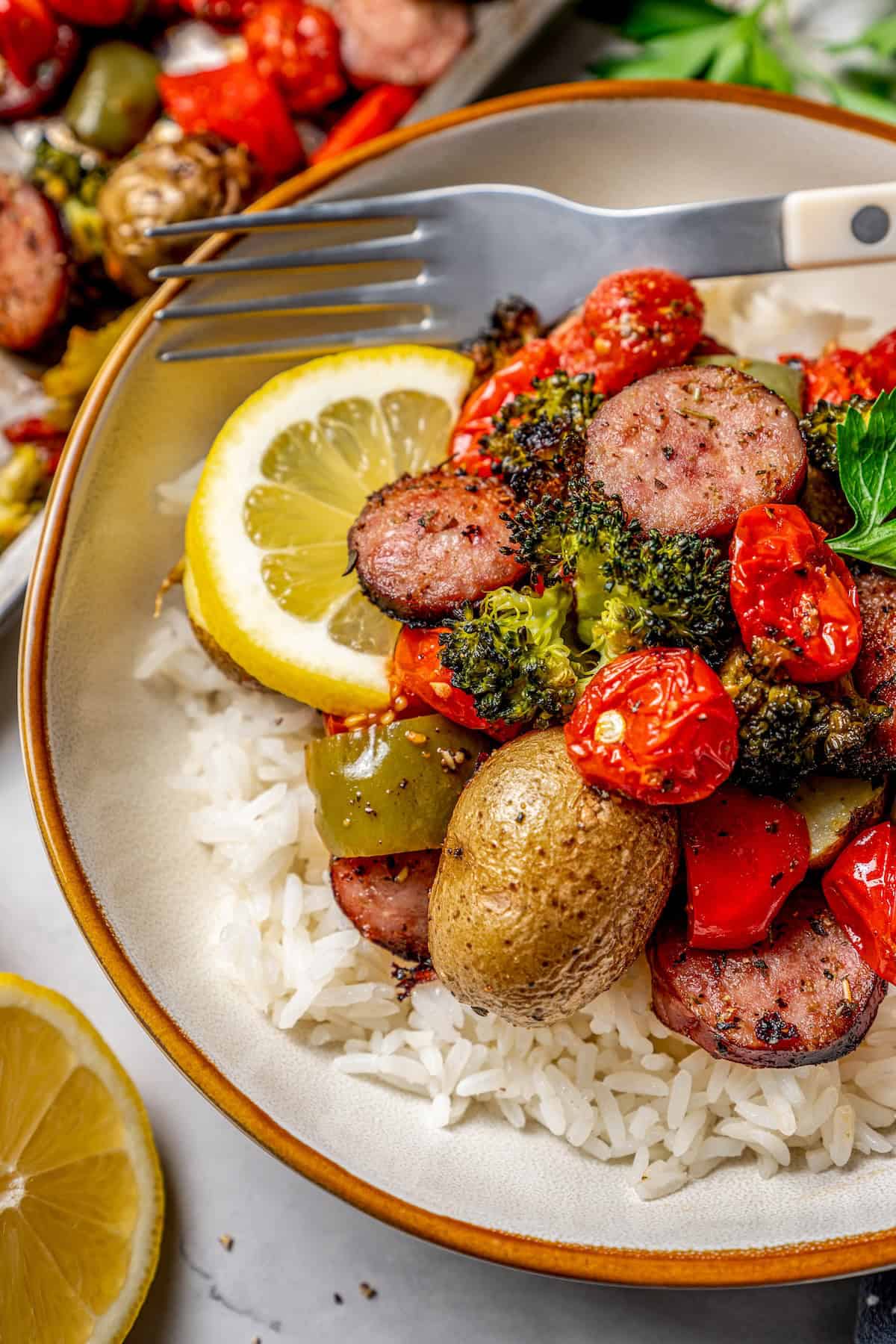 Close up of a bowl of rice topped with a mixture of sausages, veggies and potatoes alongside a sheet pan where it all roasted.