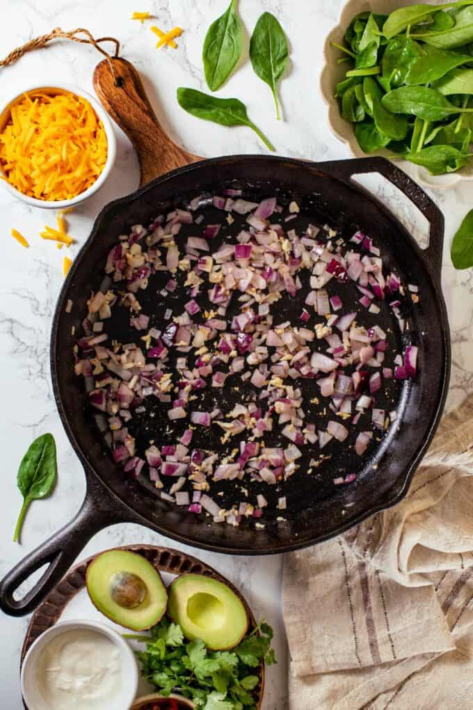 A large cast iron skillet with diced sautéed red onion and garlic.