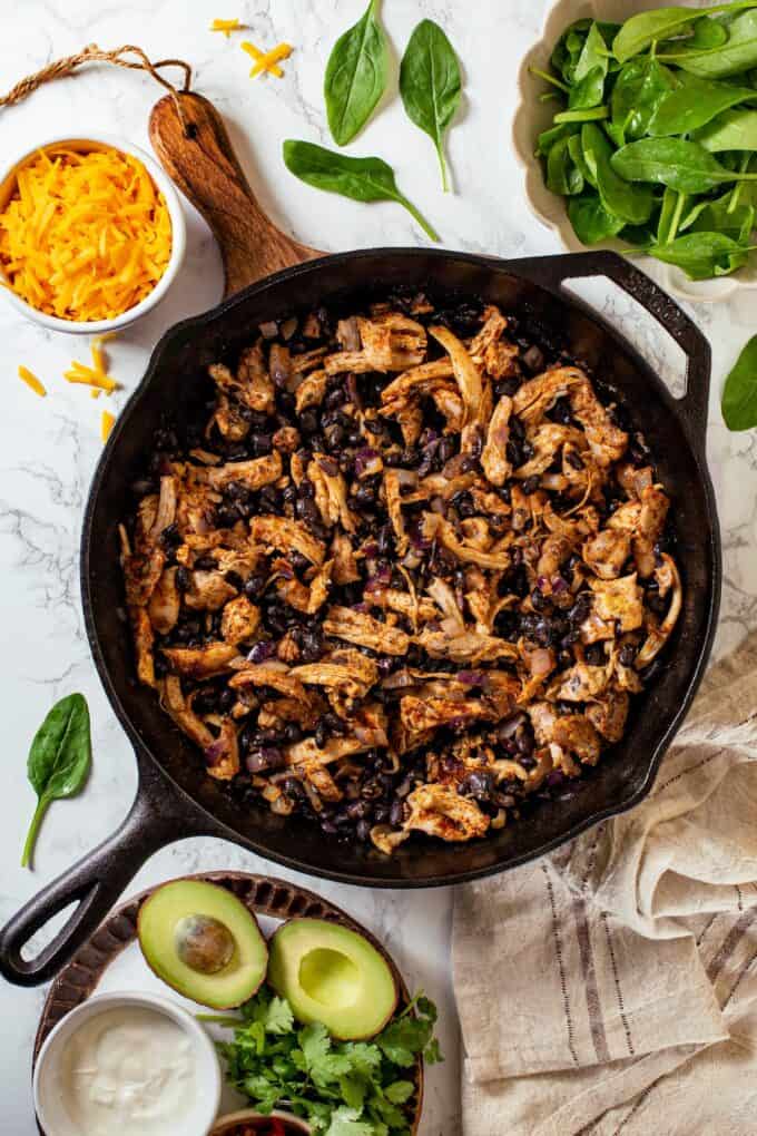 Shredded chicken and black beans in a cast iron skillet with cheese, spinach and toppings nearby.