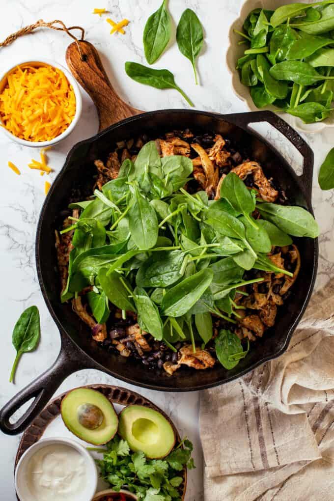 Fresh baby spinach piled on top of a chicken and black bean mixture in a cast iron skillet before being wilted down and mixed in.