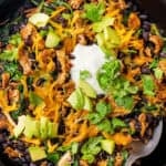 A spoon resting in a skillet with a cheesy chicken, black bean and spinach mixture topped with sour cream, avocado and cilantro and a text overlay on the photo.
