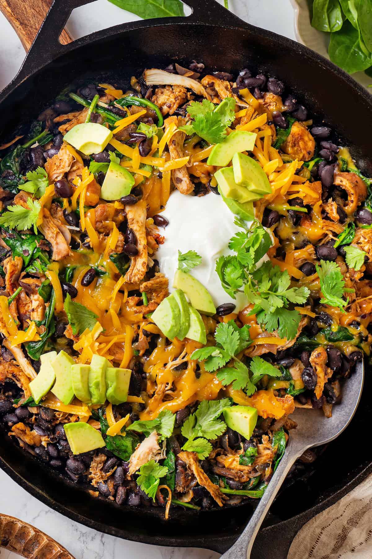 A spoon resting in a cast iron skillet full of chicken, black beans, spinach and melted cheese on top with diced avocado and fresh cilantro and sour cream as garnishes.