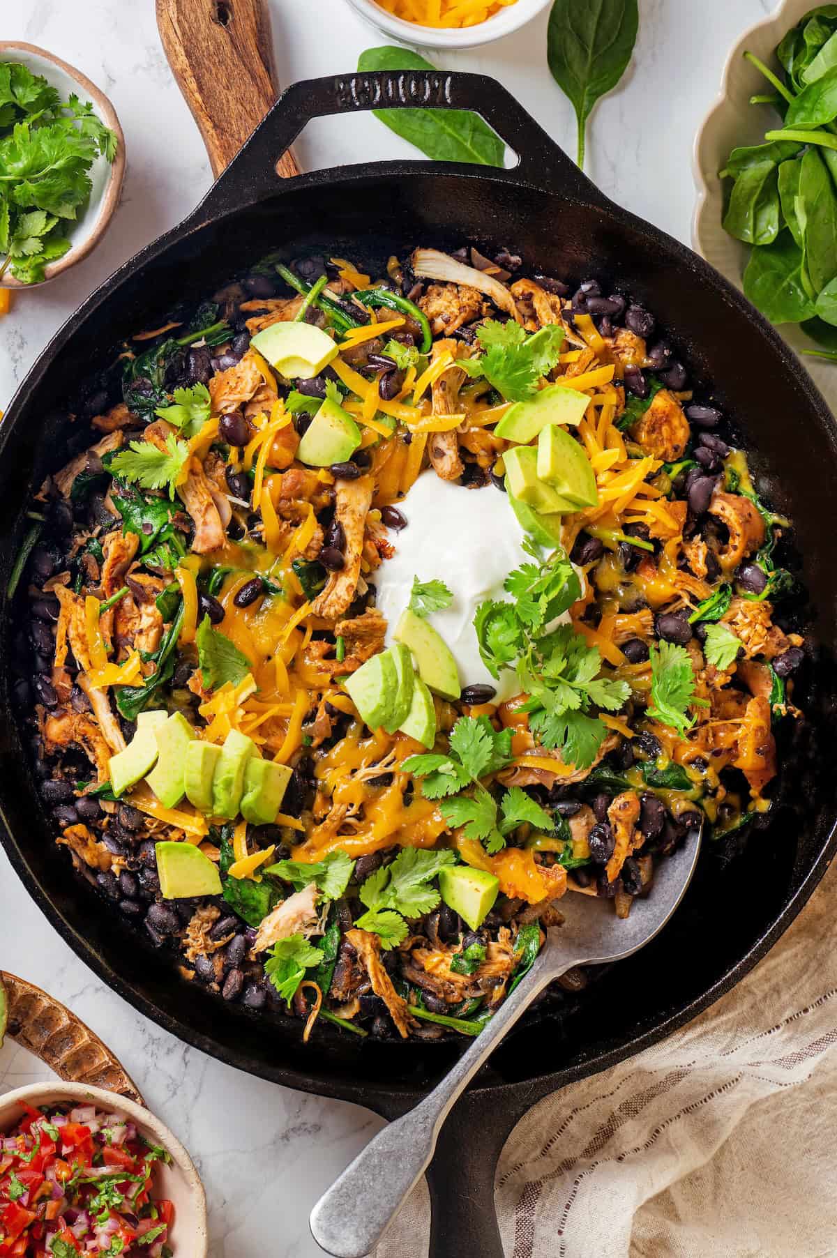 A spoon resting in a dark cast iron skillet full of chicken, black beans, spinach and melted cheese on top with diced avocado and fresh cilantro and sour cream as garnishes.