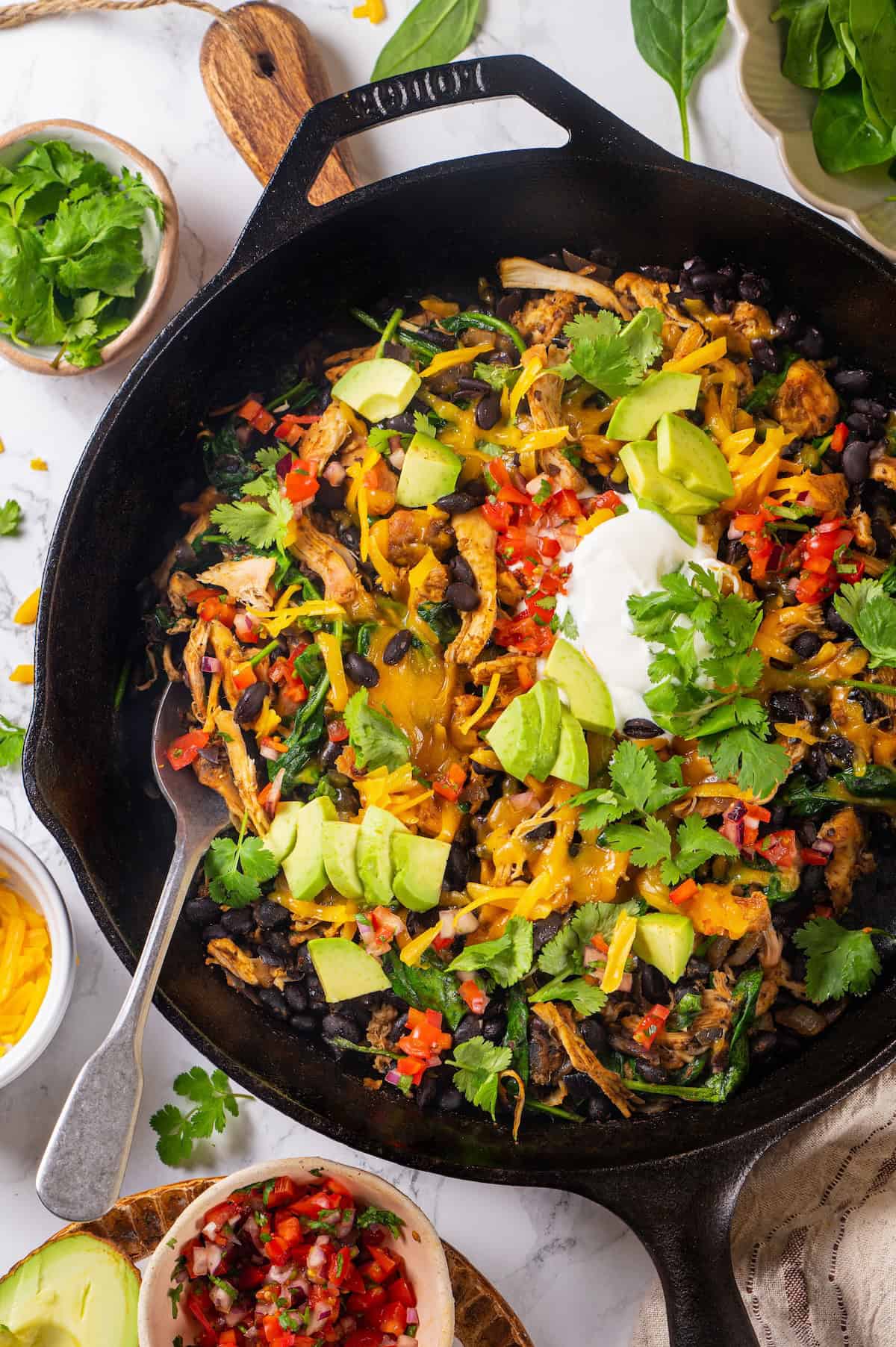 A spoon resting in a cast iron skillet full of chicken, black beans, spinach and melted cheese on top with diced avocado, salsa, fresh cilantro and sour cream as garnishes.