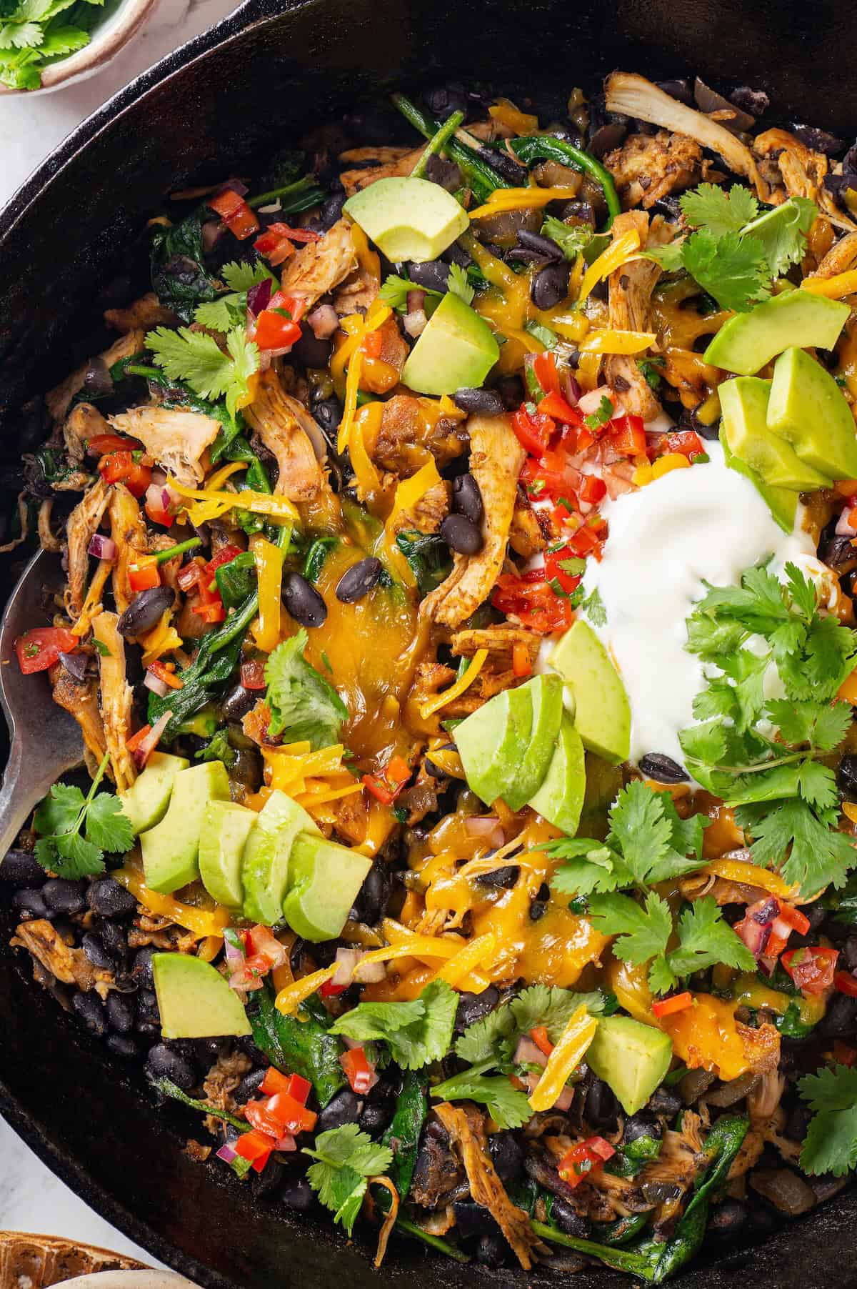 Close up of a spoon resting in a cast iron skillet full of chicken, black beans, spinach and melted cheese on top with diced avocado, salsa, fresh cilantro and sour cream as garnishes.