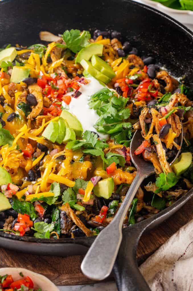 Side angle view of a spoon resting in a cast iron skillet full of chicken, black beans, spinach and melted cheese on top with diced avocado, salsa, fresh cilantro and sour cream as garnishes.