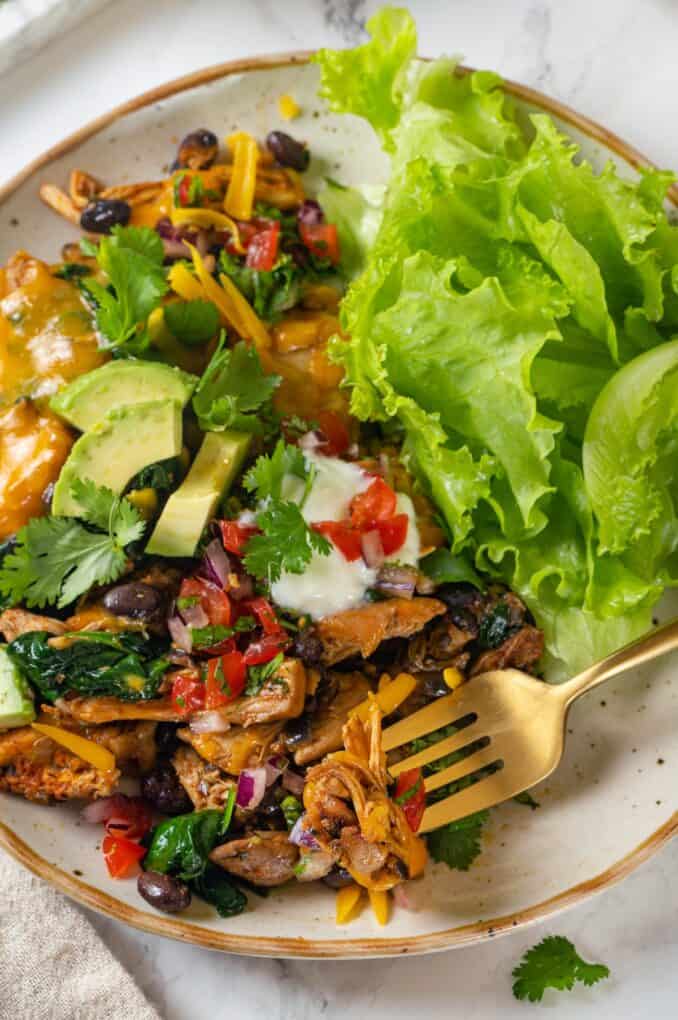 A fork resting in a bite of chicken on a plate with a cheesy chicken and black bean mixture alongside fresh leafy lettuce.