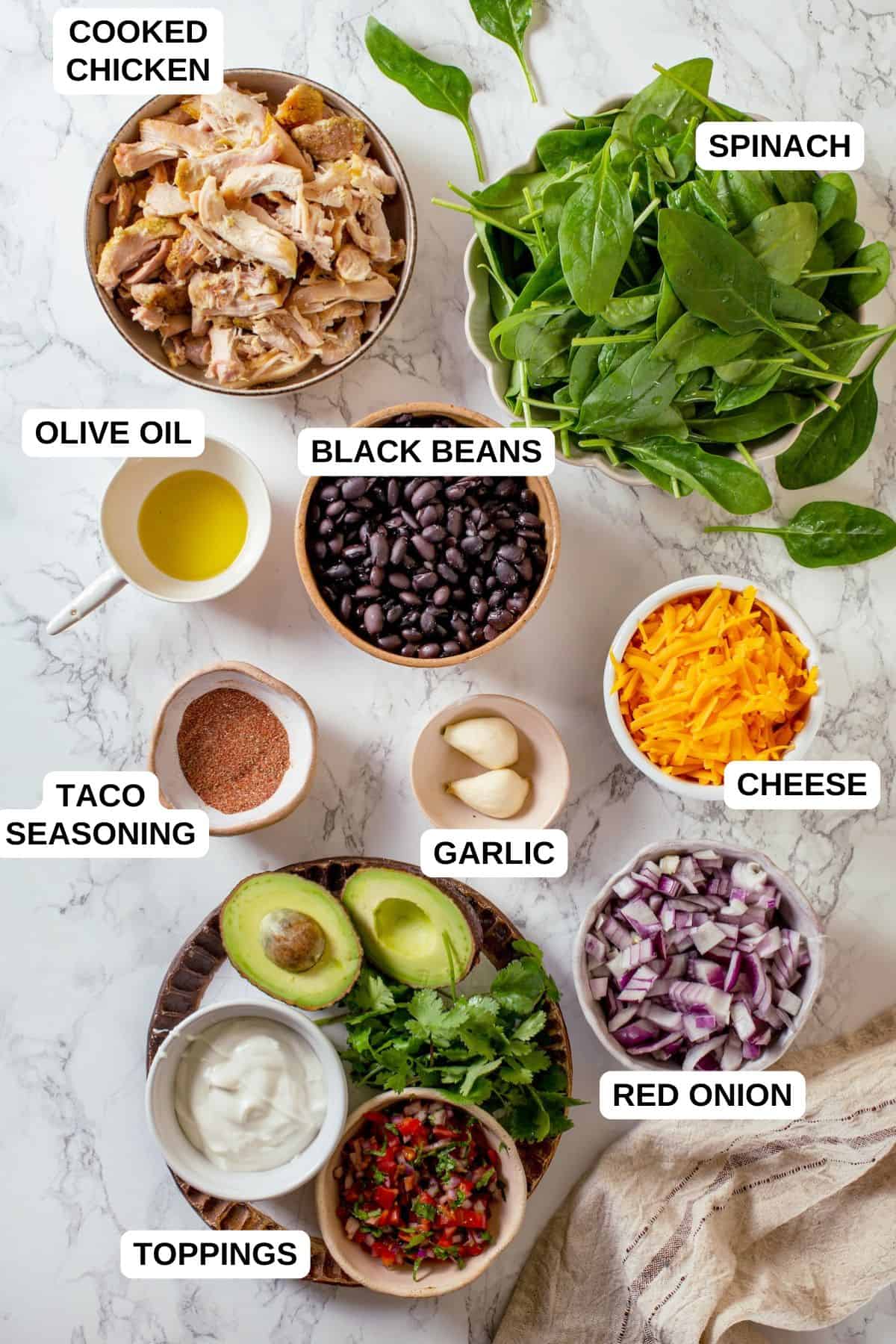 Labeled ingredients laid out on a counter in separate bowls, including cooked chicken, spinach, black beans, cheese and red onion.