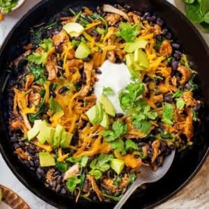 A spoon resting in a skillet with a cheesy chicken, black bean and spinach mixture topped with sour cream, avocado and cilantro.