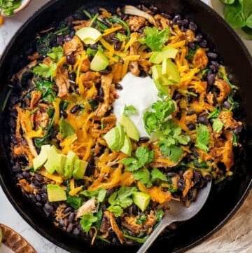 A spoon resting in a skillet with a cheesy chicken, black bean and spinach mixture topped with sour cream, avocado and cilantro.