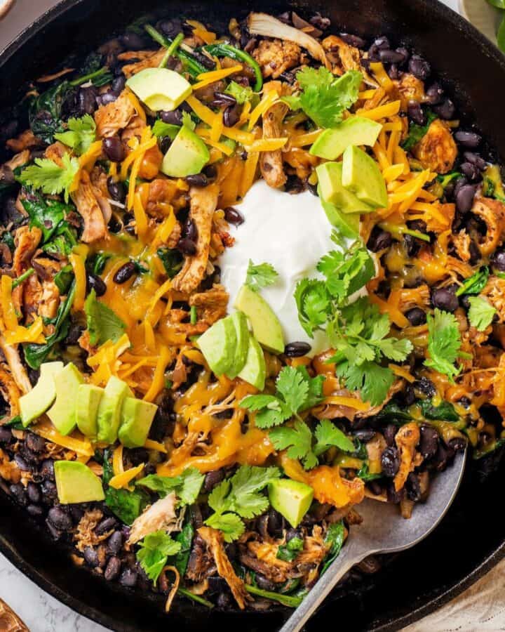 A spoon resting in a skillet with a cheesy chicken, black bean and spinach mixture topped with sour cream, avocado and cilantro.