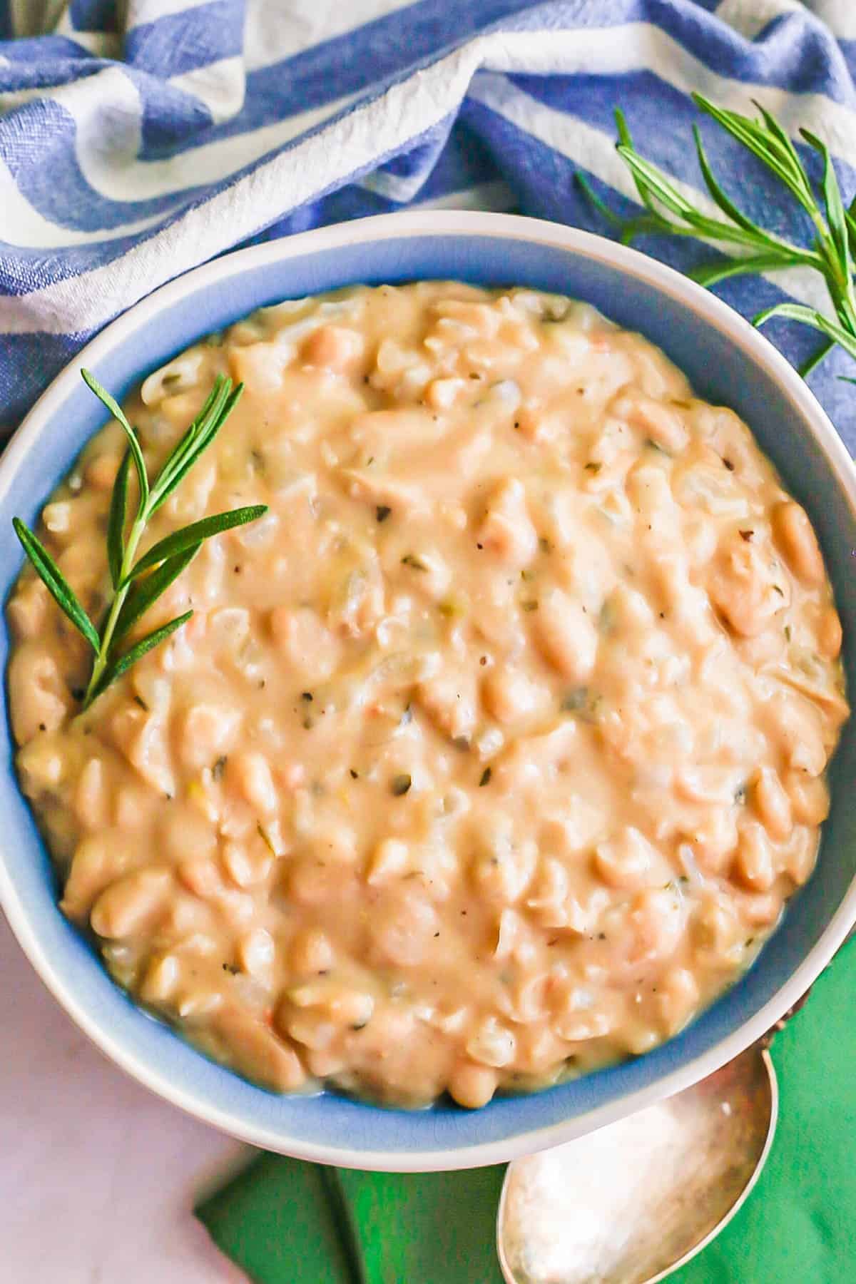 A large blue bowl full of creamy white beans with sprigs of rosemary on top and to the side.