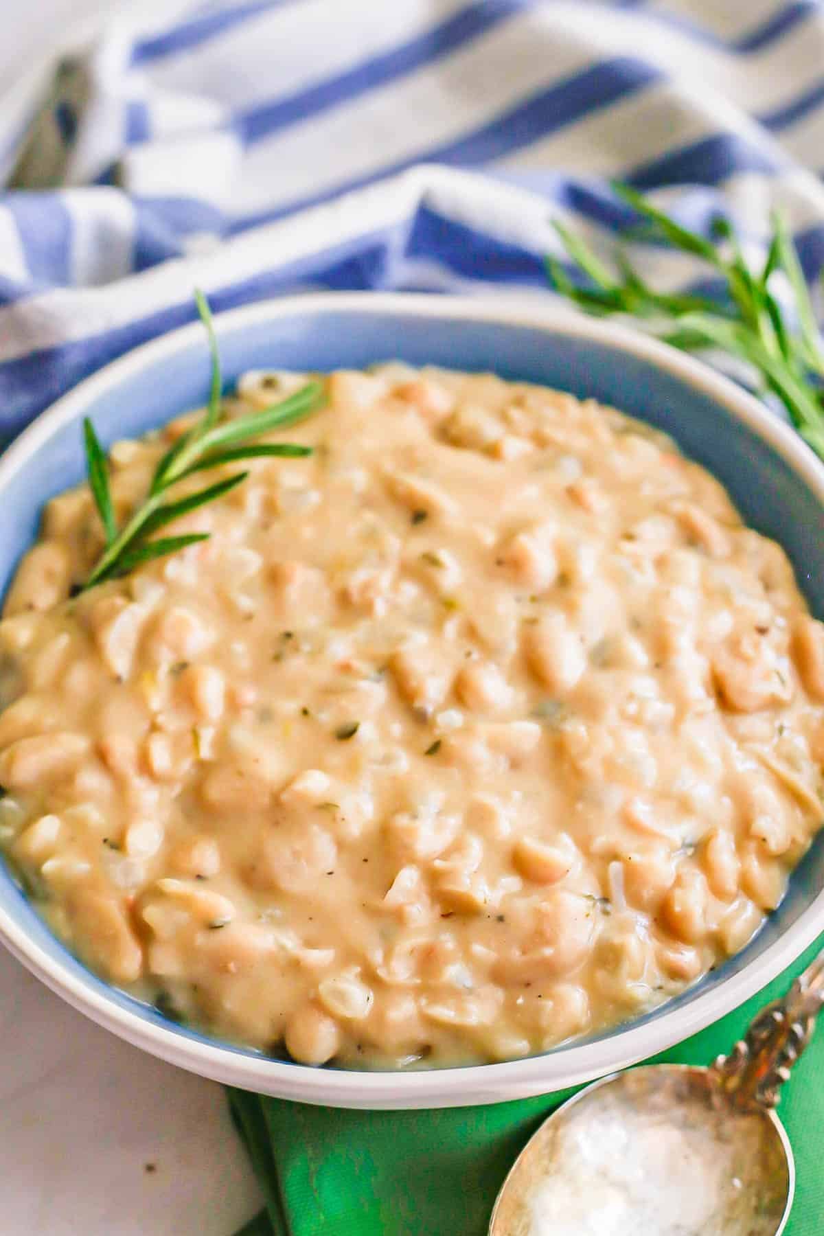 A blue bowl full of creamy white beans with sprigs of rosemary on top and to the side with a spoon to the side.