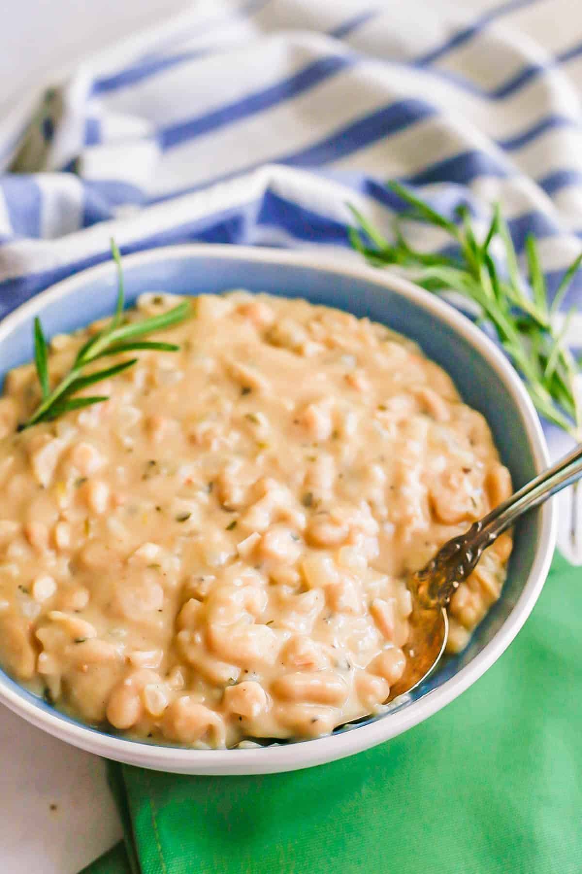 A spoon resting in a blue and white serving bowl of creamy white beans and onions with rosemary on top.
