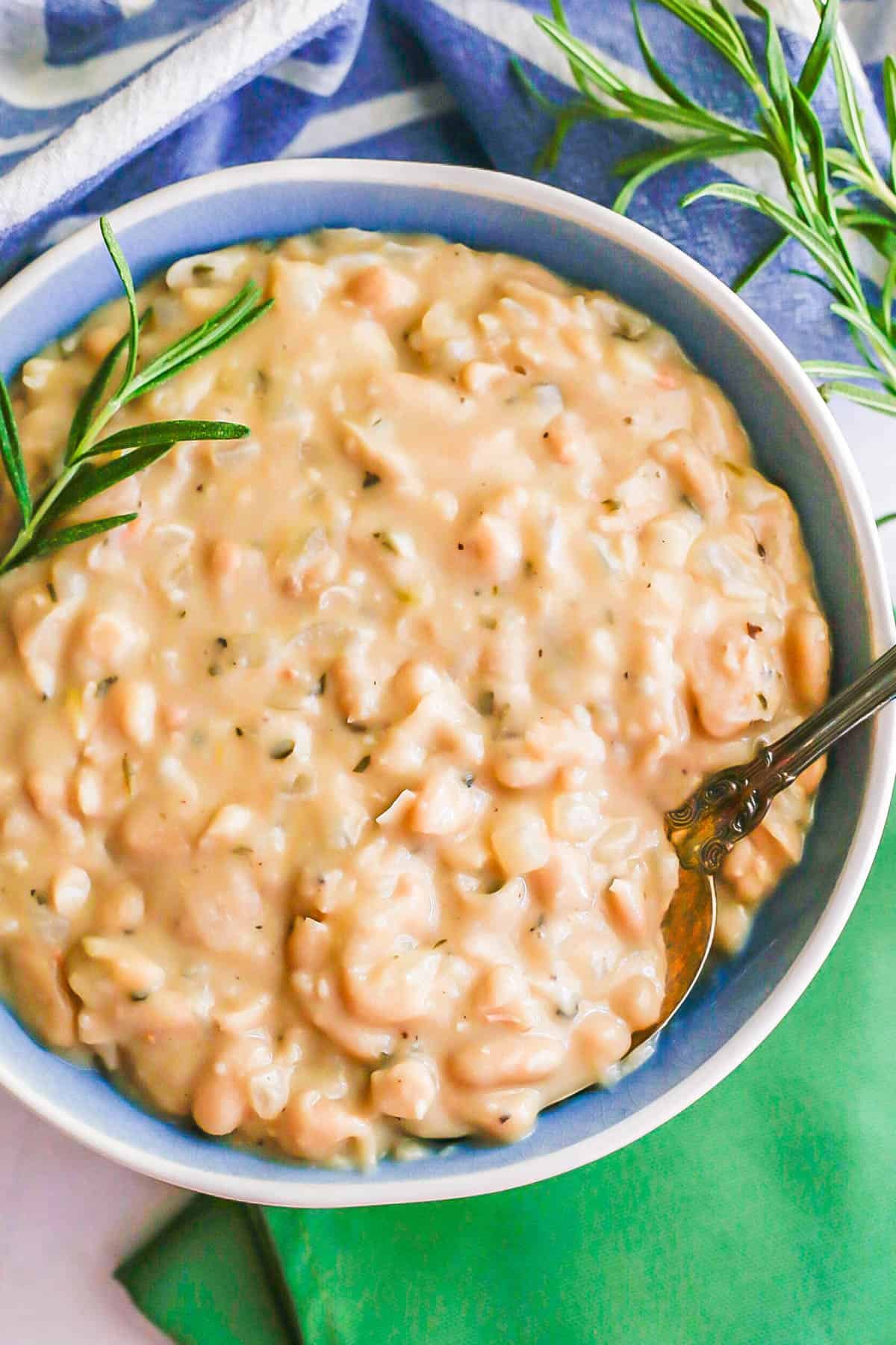 A spoon resting in a blue and white serving bowl of creamy white beans and onions with a sprig of rosemary on top.