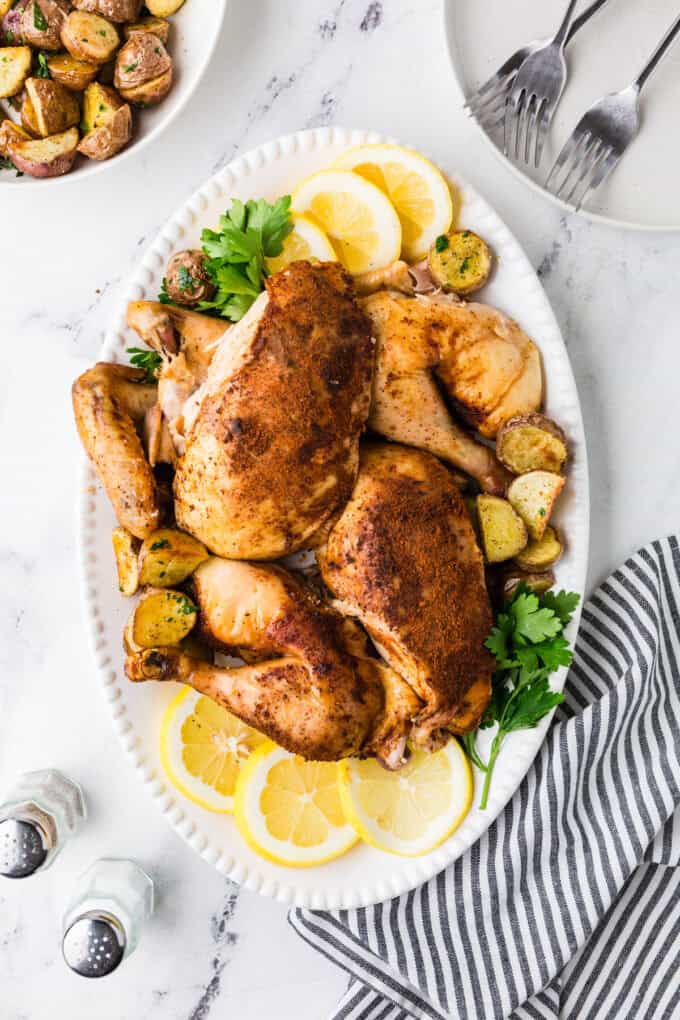 A carved whole chicken served on a large white platter with lemon wedges, parsley and roasted potatoes.