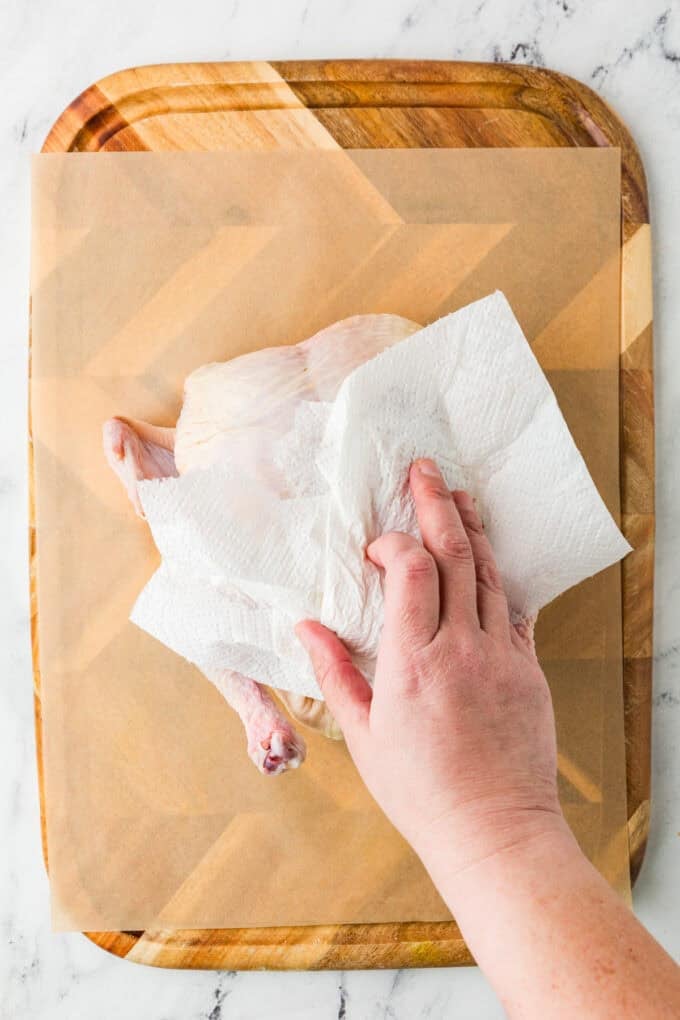A hand with paper towels patting dry a whole chicken on a cutting board.