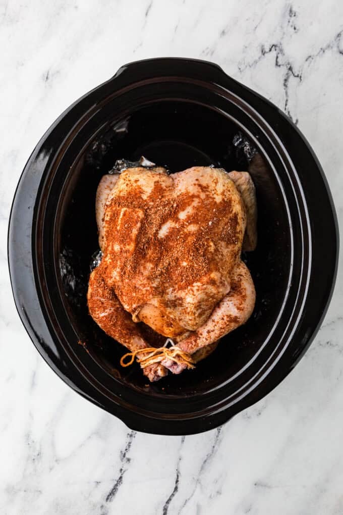 A seasoned and trussed whole chicken in a crock pot insert before being cooked.