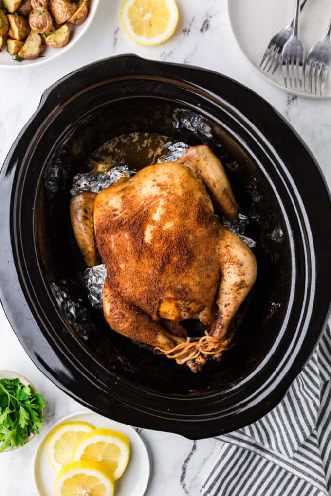 A seasoned and trussed whole chicken in a crock pot insert after being cooked.