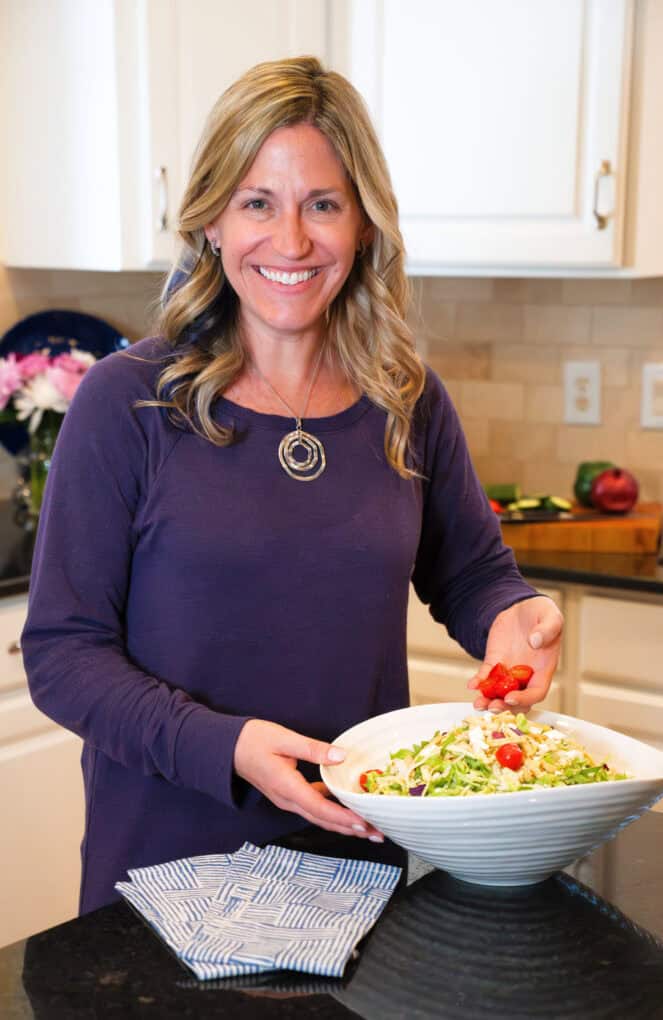 A woman in a kitchen sprinkled tomatoes onto a salad in a large white bowl.