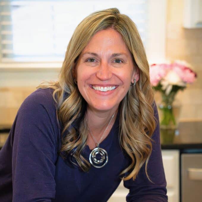 A woman in the kitchen leaning on a counter with flowers in the background.