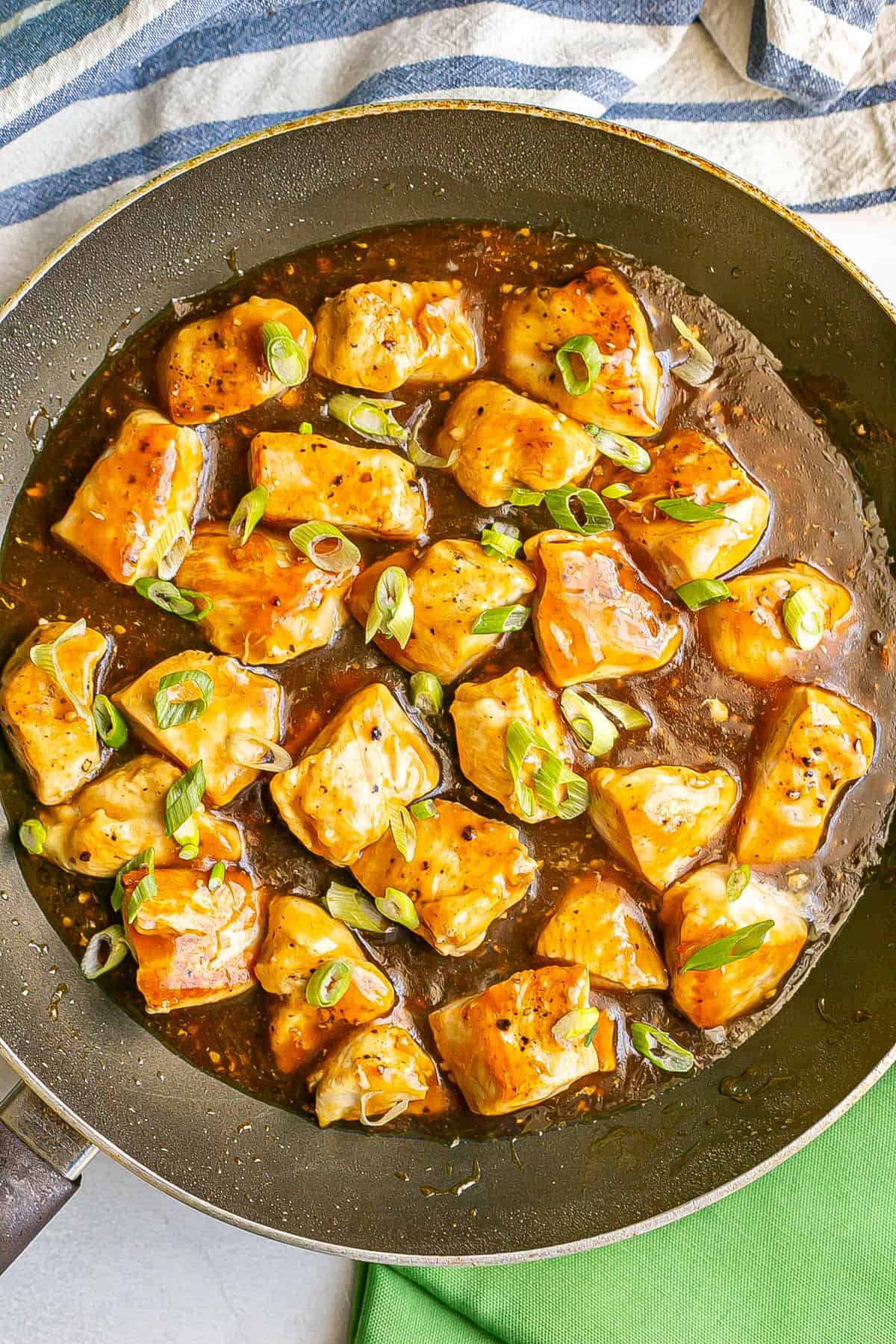 Seared cubed pieces of chicken in a teriyaki sauce in a large skillet with sliced green onions sprinkled on top.