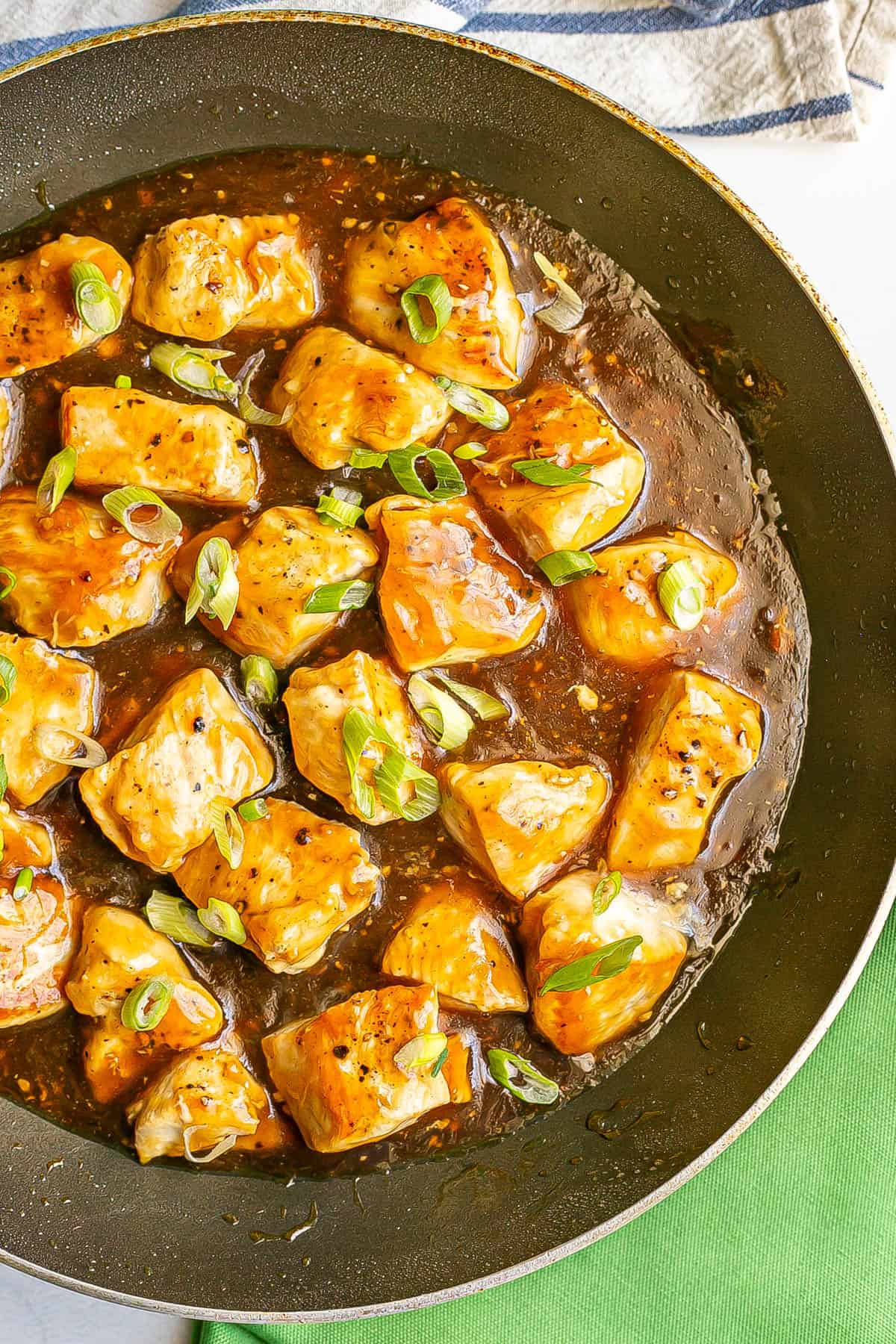 Close up of seared cubed pieces of chicken in a teriyaki sauce in a large skillet with sliced green onions sprinkled on top.