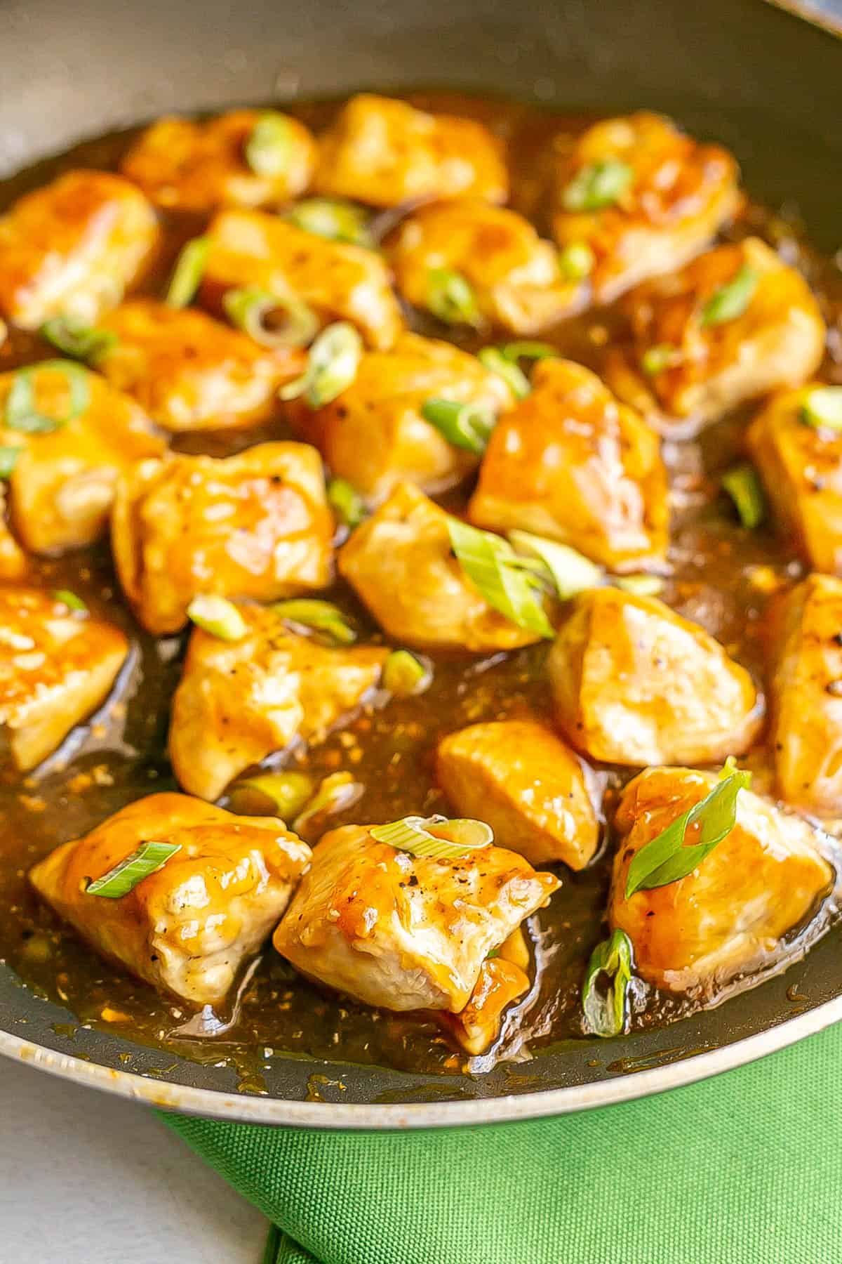 Seared cubed pieces of chicken in a teriyaki sauce in a large skillet with sliced green onions sprinkled on top and a green napkin to the side.