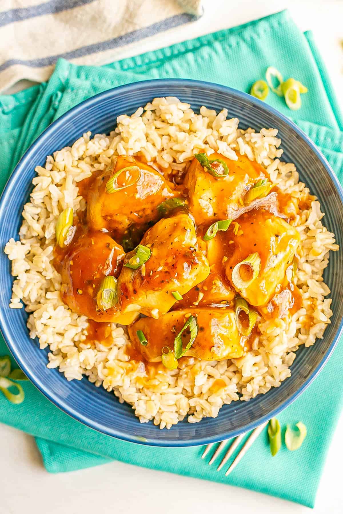 Teriyaki chicken pieces served over steamed brown rice in a blue bowl with sliced green onions sprinkled on top.