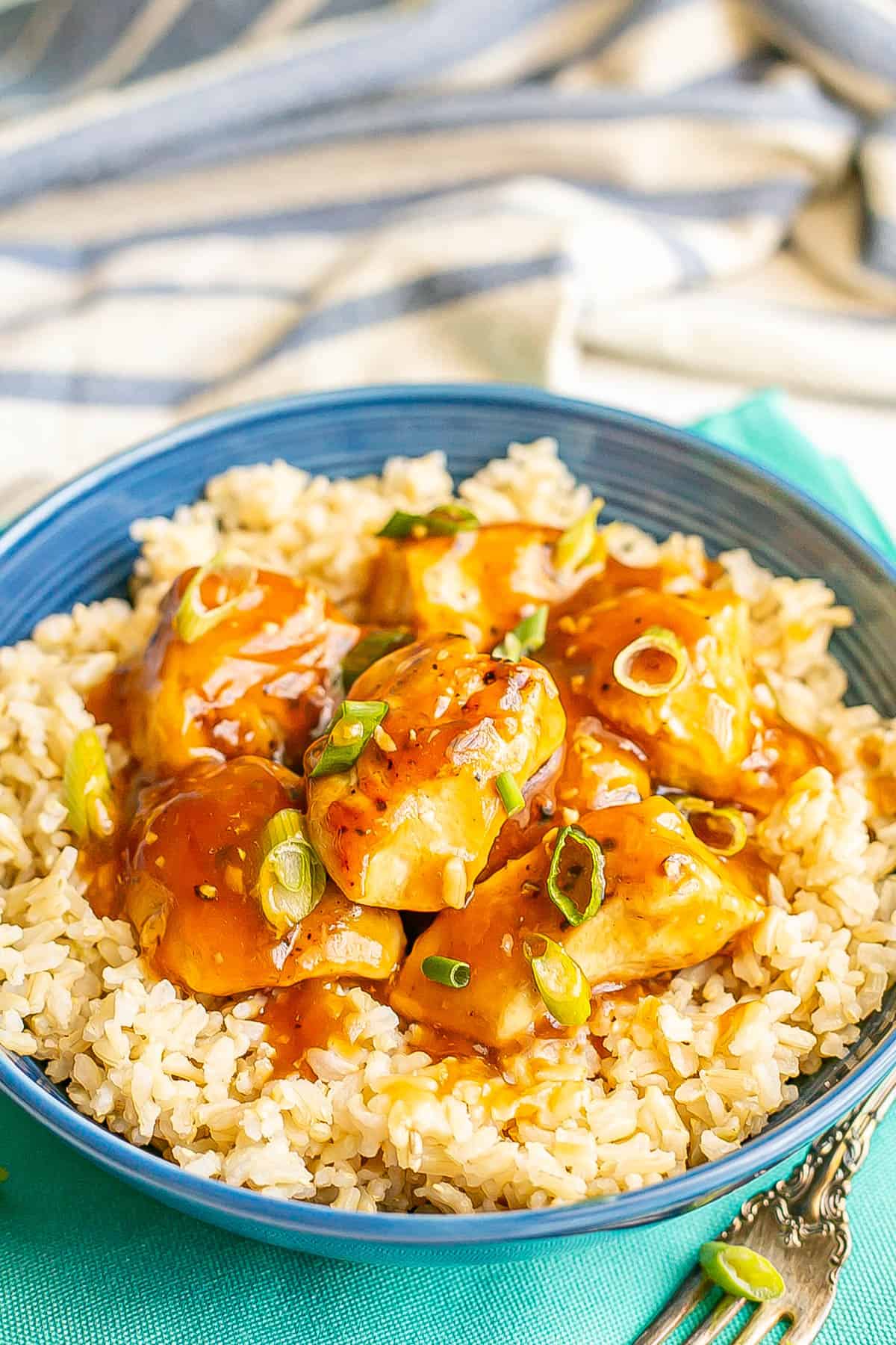 Teriyaki chicken pieces and sauce served over steamed brown rice in a blue bowl with sliced green onions sprinkled on top.