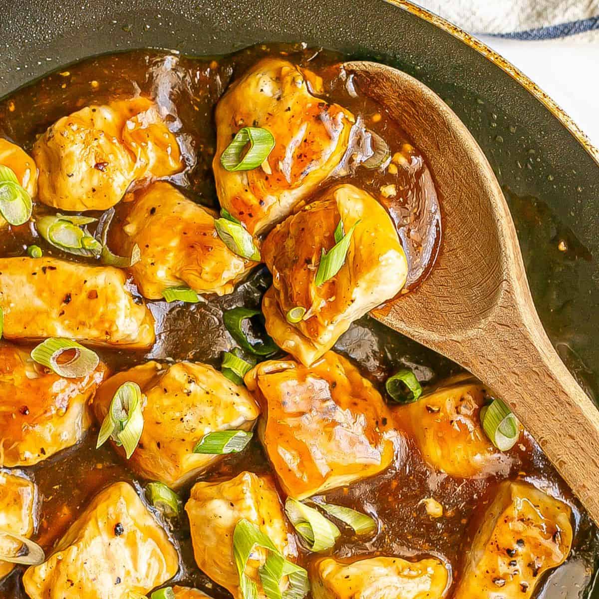 Close up of a wooden spoon resting in a skillet with cubed chicken pieces in a teriyaki sauce with sliced green onions on top.