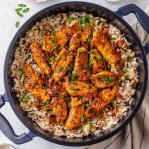 Seasoned cooked chicken strips atop a large dark pot of rice with black eyed peas topped with sliced green onions and cooked, crumbled bacon.