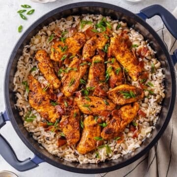 Seasoned cooked chicken strips atop a large dark pot of rice with black eyed peas topped with sliced green onions and cooked, crumbled bacon.