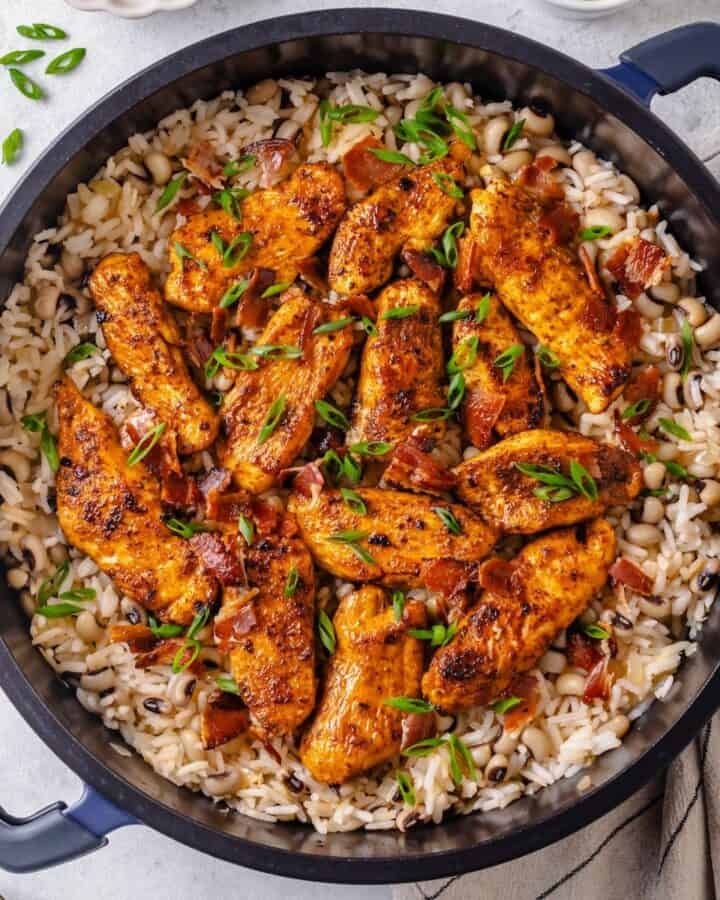 Seasoned cooked chicken strips atop a large dark pot of rice with black eyed peas topped with sliced green onions and cooked, crumbled bacon.