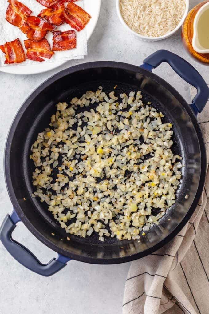 Sautéed onion and garlic in a large dark pot with cooked bacon on a plate to the side.