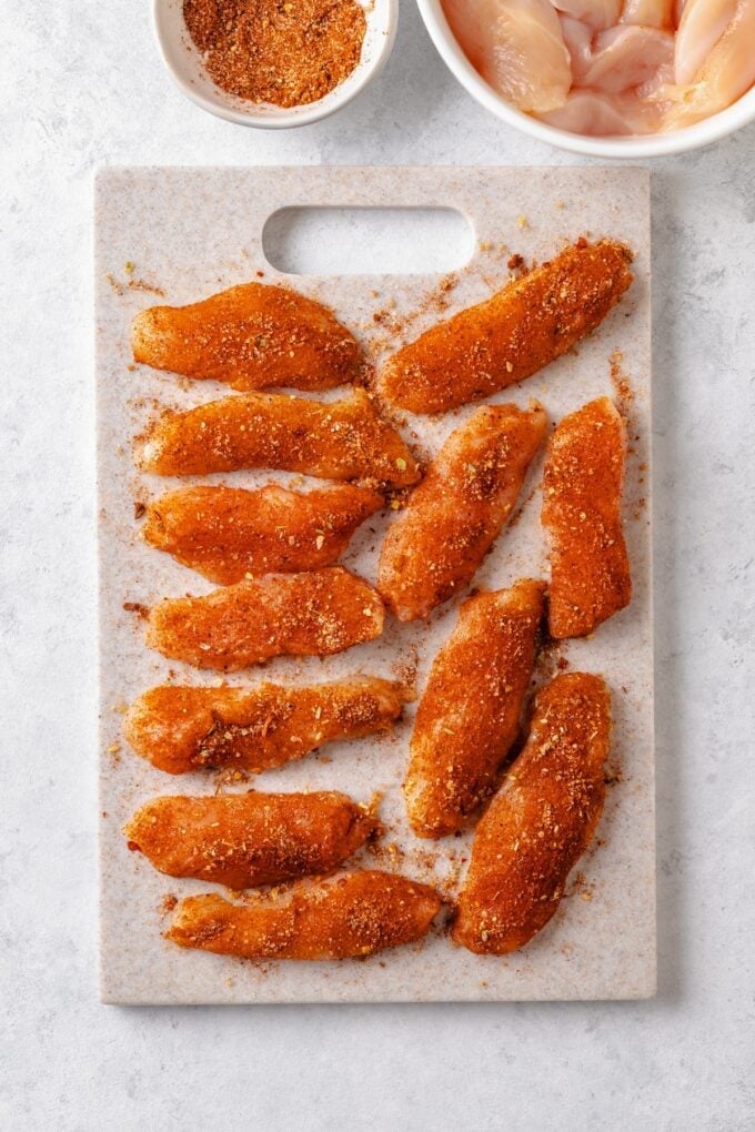 Strips of chicken breasts on a cutting board seasoned with a spice mixture.
